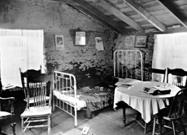 Fairdale 1932 - Elling Ohnstad's sod house in Fairdale on June 24, 1932. The windows adorned with curtains, the patch-work quilt on the metal framed bed, and the round table complete with tablecloth, all demonstrate some of the warm comforts contained within a sod house. Pictures and a calendar hanging on the wall complete the scene.